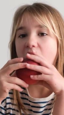 Renkli çizgili gömlek giymiş küçük bir kız açık havada güneşli bir günün tadını çıkarırken mutlu bir şekilde kırmızı elma yiyor.