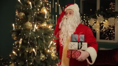 Noel Baba, güzel dekore edilmiş bir Noel ağacının önünde elinde renkli hediyeler ve hediyelerle neşeli bir şekilde duruyor.