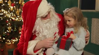 Bu iç açıcı sahnede, küçük bir kız Noel Baba 'yla neşeyle etkileşime geçiyor, elinde şeker kamışı ve bir hediye, gözleri heyecandan parlıyor.