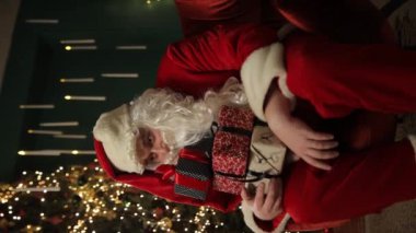 Noel Baba kostümü giymiş bir adam süslü bir Noel ağacının yanında elinde neşeyle renkli hediyeler tutuyor ve tatil sezonunun özünü somutlaştırıyor.