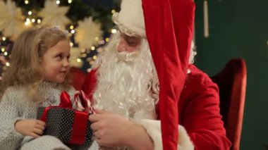 Noel Baba 'nın kucağında sevinçle oturan küçük bir kız, güzel bir hediye paketi tutuyor, heyecanla Noel gününü bekliyor.