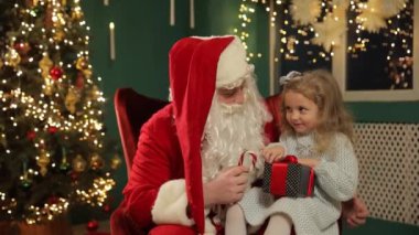 Küçük bir kız elinde baston şekerle Noel Baba 'nın kucağına sevinçle oturur. Güzel dekore edilmiş bir Noel ağacı şenlikli atmosferi güçlendirir.