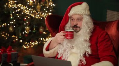 Bu tuhaf tatil videosunda, Noel Baba rahat bir ortamda kahve içerken dizüstü bilgisayar ve telefonla birden fazla iş yapıyor.