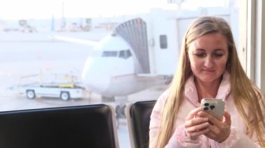Bu videoda, havaalanında bir kadın cep telefonuna dalmış, beklentinin ve modern bağlantının karışımını vurguluyor.