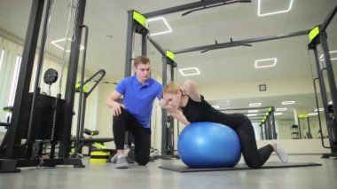 Bir adam pilates topuyla bir kadına yardım ediyor, bacağına ve bacak kaslarına odaklanıyor, spor kıyafetlerini spor salonunda giyip spor salonlarında spor yapıyor. Dikey video