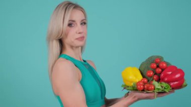 Neşeli bir kadın mavi arka planda taze sebzeler sunar. Gülümsemesi pozitiflik saçıyor, sağlıklı yiyecekleri ve sağlıklı yaşam tarzını ön plana çıkaran renkli bir çeşitlilik sergiliyor.