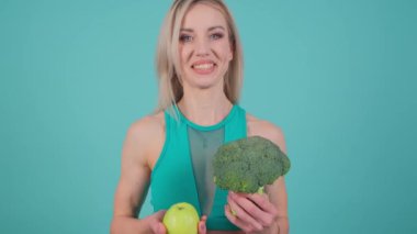 Sağlıklı yaşam tarzını destekleyen bir kadın ve arka planda taze ürünler. Elinde brokoli ve elmayla besleyici yiyecekler sunuyor..