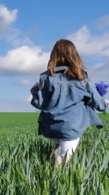 Şık bir kot ceket giyen genç bir kız mavi gökyüzünün altındaki güzel yeşil bir tarlada mutlu mesut dolaşır..