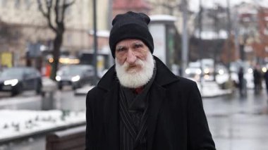 Uzun sakallı, sıcak kış kıyafetleri giymiş yaşlı bir adam karlı bir şehir ortamında geziniyor..