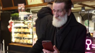 Uzun beyaz sakallı yaşlı bir adam rahat rahat oturuyor ve rahat bir kafe ortamında telefonunu kullanıyor.