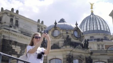 Güneşli güzel bir günde, genç bir kadın arka planda tarihi bir bina bulunan bir selfie çekiyor.