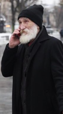 Sıcak kışlık ceket ve şapka giymiş yaşlı bir adam dışarıda akıllı telefonuyla mutlu bir şekilde konuşuyor..
