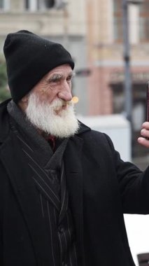 Şehir ortamında, beyaz sakallı, koyu renk elbiseli yaşlı bir adam coşkuyla selfie çeker.