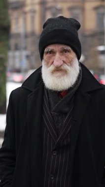 Zarif siyah takım elbiseli, uzun beyaz sakallı yaşlı bir adam karlı bir şehrin ortasında duruyor ve işaret parmağını sallıyor..