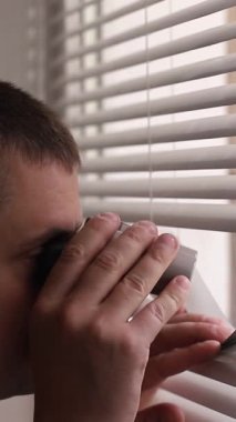 Bir adam dikkatle pencerenin perdelerinden dışarı bakıyor, sadece merakı değil, aynı zamanda tetikte olma duygusunu da ortaya çıkarıyor.