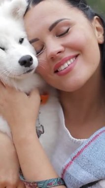 Neşeli bir kadın, tüylü bir Samoyed köpeğini kucaklıyor ve kucaklıyor. Karşılıklı sevgilerini ve sıcaklığını güzelce sergiliyor..