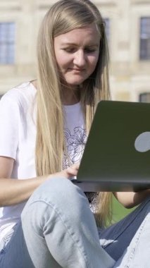 Odaklanmış bir kadın, çarpıcı tarihi binalarla çevrili çimlerin üzerinde otururken dizüstü bilgisayarı üzerinde çalışıyor. Dikey video.