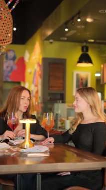 İki kadın neşeli bir şekilde içkilerini ve kahkahalarını renkli sanatlarla çevrili canlı bir restoranda paylaşıyor.