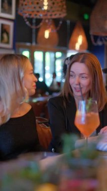 İki kadın şık bir restoran ortamında lezzetli içkiler eşliğinde neşeli ve ilgi çekici bir sohbetin tadını çıkarıyorlar.