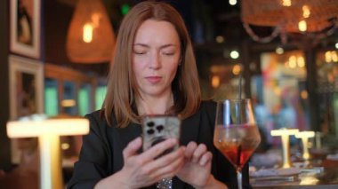 Odaklanmış bir kadın şık bir barda akıllı telefonunu kullanıyor. İçkisinin ve modern atmosferin tadını çıkarıyor.