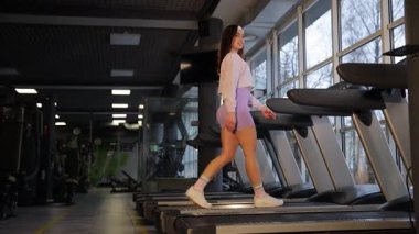 Genç bir kadın, sağlıklı bir yaşam tarzını destekleyen parlak ve modern bir spor salonundaki koşu bandında güvenle yürüyor.