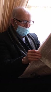 Koruyucu maske takan yaşlı bir adam bugün evdeyken gazete okumaya dalmış durumda.