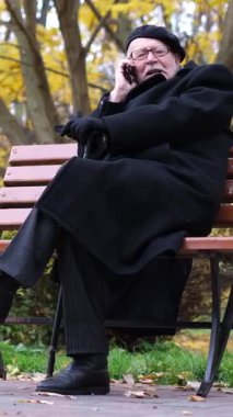 Yaşlı bir adam, şık siyah bir ceket giyiyor, güzel bir park manzarası izlerken bir telefon görüşmesi yapıyor..