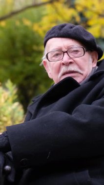 Şık bir bere takan yaşlı bir beyefendi düşünceli ve düşünceli bir şekilde canlı bir sonbahar yeşilliğinin ortasında oturur..