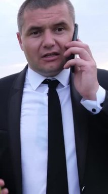 Keskin giyimli bir işadamı dışarıda duruyor, enerjik bir şekilde telefonla konuşuyor, aciliyet ve profesyonellik gösteriyor.