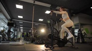 Şık giyinen formalı genç bir kadın sofistike bir spor salonunda eliptik bir makine kullanıyor.