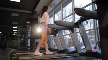 Modern bir spor salonundaki koşu bandında yürüyen genç bir kadın zindelik ve sağlığın önemini vurguluyor..