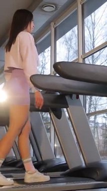 Modern bir spor salonunda koşu bandında yürüyen bir kadın aktif kalır. Dikey video.