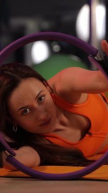 Kendini işine adamış genç bir kadın pilates ringi ile paspas egzersizleri yapıyor ve spor yapmaya olan bağlılığını gösteriyor. Dikey video.