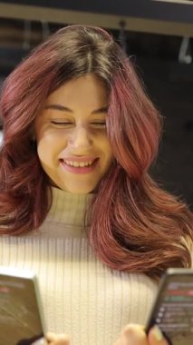 Pembe saçlı genç bir kadın, bir elektronik mağazasından hangisini alacağına karar veren iki telefonu karşılaştırıyor. Dikey video.