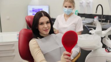 Neşeli bir hasta, parlak bir gülümseme takınarak dişçi koltuğunda oturuyor, diş muayenesi sırasında elinde bir ayna tutuyor.