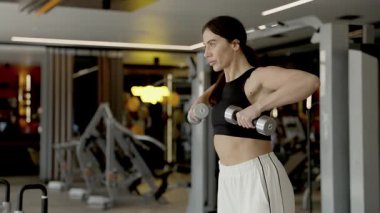 Kararlı bir kadın, modern bir spor salonunda dambıllarla güçlü bir antrenman yapıyor.