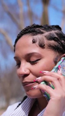 Telefonunu açık havada mutlu bir şekilde kullanan, örgülü saç stili ve şık tırnaklarını sergileyen bir kadının yakın çekimi.