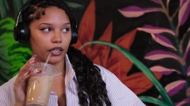 Afro-Amerikalı bir kadın kulaklık takıyor ve ferahlatıcı buzlu kahvenin tadını çıkarıyor.