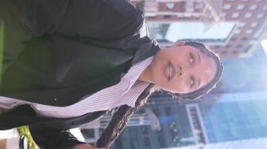 Afro-Amerikalı bir kadın, güneş ışığıyla aydınlanan canlı bir şehir ortamında kendine güvenerek duruyor..