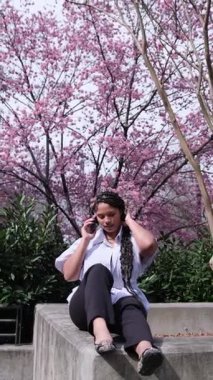 Etrafı güzel kiraz çiçekleriyle çevriliyken gündelik kıyafetlerini giymiş bir kadın telefonda zarif bir şekilde konuşur..