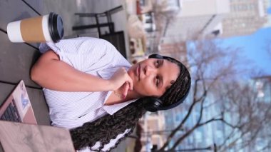 Kulaklık takan, kahvesini yudumlayan bir kadın, dışarıda canlı bir şehir ortamında otururken derin düşüncelere dalıyor.