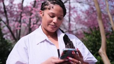 Genç Amerikalı kadın ağaçlarla çevrili güzel bir parkta otururken akıllı telefonuyla mutlu mesut eğleniyor..