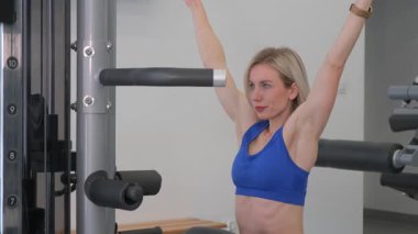 Spor tişörtlü güzel sarışın kadın modern bir egzersiz makinesinde kollarını kaldırıyor. Kol kasları için egzersiz.