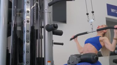 Spor tişörtlü güzel sarışın kadın modern bir egzersiz makinesinde kollarını kaldırıyor. Kol kasları için egzersiz.