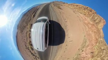 Panoramik manzara, açık mavi bir gökyüzünün altında açık bir çöl yolunda giden bir arabayı gösteriyor..
