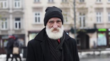 Bilge yaşlı bir adam işlek bir şehir caddesinde duruyor, kış soğuğuna karşı direncini gösteriyor.