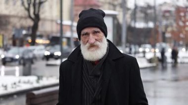 Etkileyici sakallı ve kışlık giysileri olan yaşlı bir adam enerji dolu kalabalık bir şehir caddesinde duruyor.