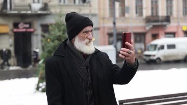 Siyah ceketli, beyaz sakallı ve bereli yaşlı bir adam şehirde neşeyle karlı bir selfie çekiyor.