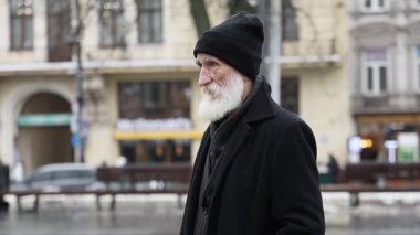 Uzun sakallı ve örgü şapkalı yaşlı bir adam karlı bir şehir manzarasında düşünceli bir şekilde duruyor..