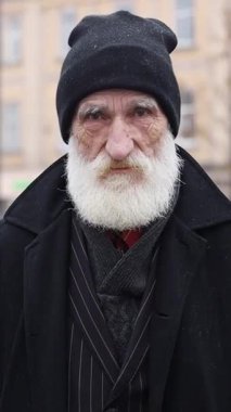 Asil, beyaz sakallı, şehir ortamında siyah paltolu ve şapkalı yaşlı bir adam..